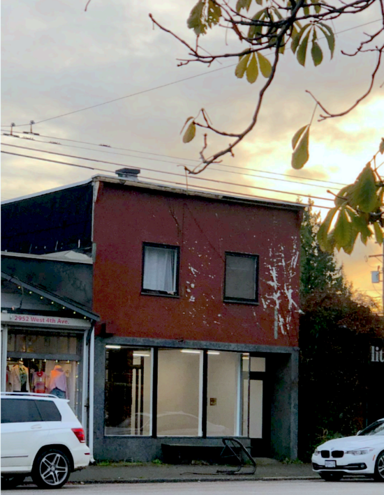 A burgundy 2-story building with large street-level windows. A sunset is shinign through the clouds in the background, and some tree branches are in the foreground.